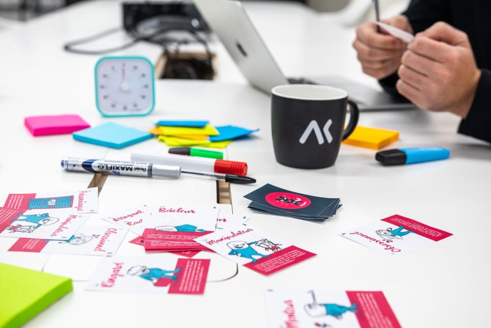 Table comprenant des posts-it, une tasse, un timer et des cartes du jeu. On voit les mains d'une personne tenant une carte